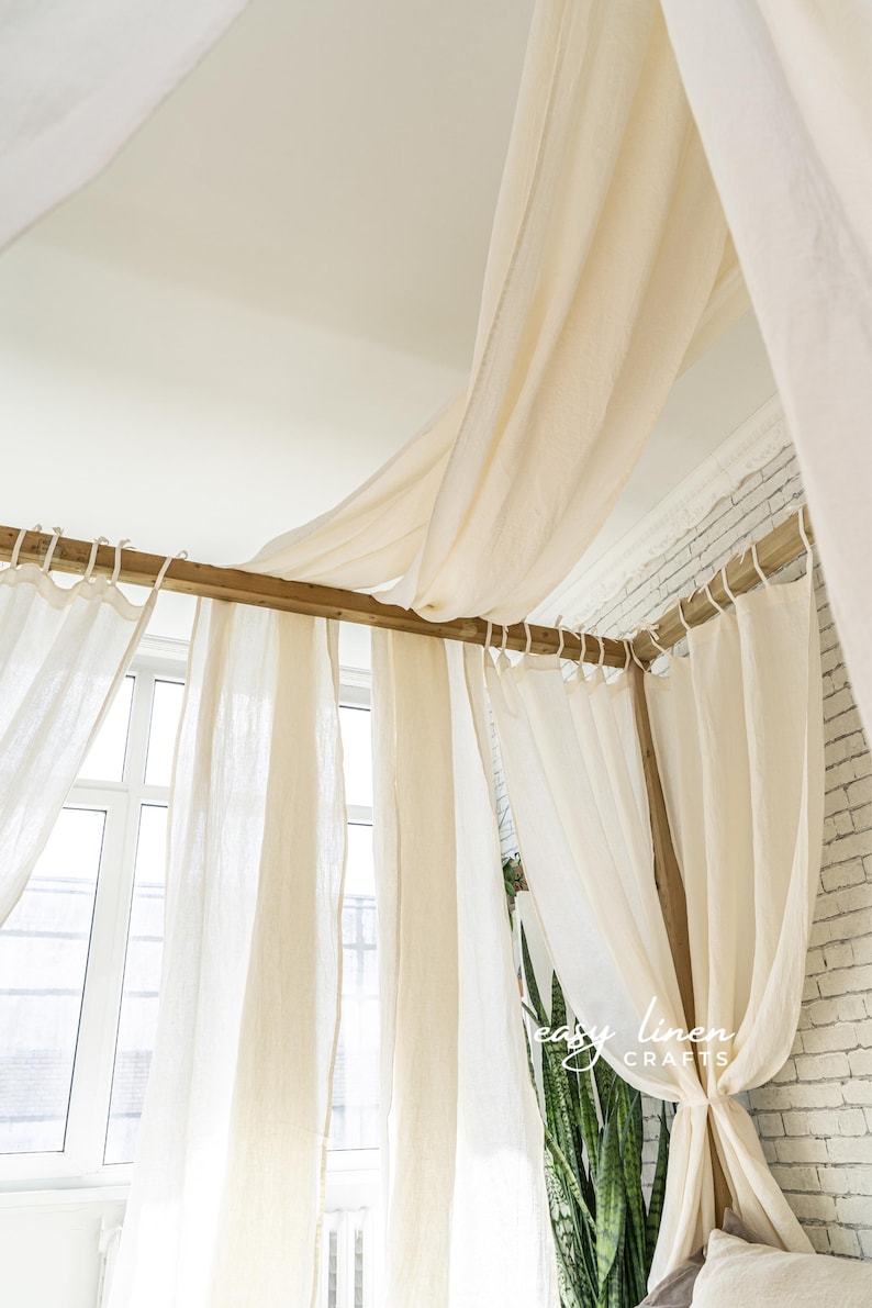 May include: A white linen canopy bed with a wooden frame and white curtains. The bed is decorated with a green plant and a white pillow.