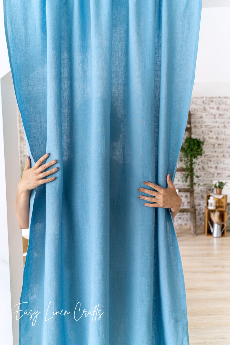 May include: A light blue linen curtain with a subtle texture. The curtain is hanging in a room with a wooden floor and a brick wall in the background.