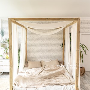 May include: A wooden canopy bed frame with a white linen canopy and a beige linen duvet. The bed is sitting on a wooden platform and has a white and beige striped rug in front of it.
