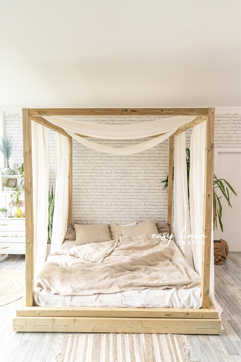 May include: A wooden canopy bed frame with a white linen canopy and a beige linen duvet. The bed is sitting on a wooden platform and has a white and beige striped rug in front of it.