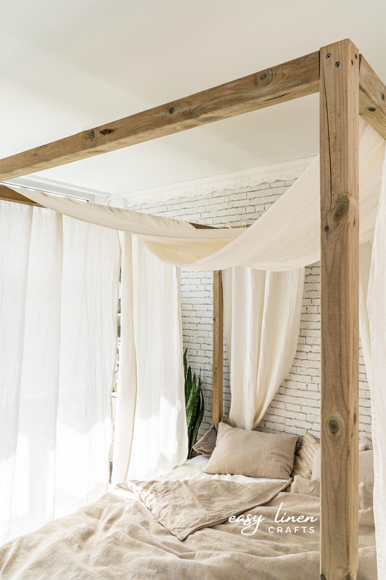 May include: A wooden canopy bed frame with white linen curtains and a beige linen duvet and pillows. The bed is in a room with a white brick wall.