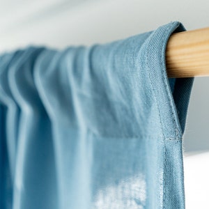 May include: Close-up of a light blue curtain hanging from a wooden rod. The curtain has a subtle texture and a stitched edge.