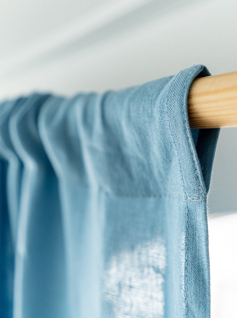 May include: Close-up of a light blue curtain hanging from a wooden rod. The curtain has a subtle texture and a stitched edge.