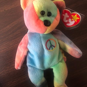 May include: A tie-dye blue, green, yellow, orange, and pink beanie baby with a peace sign on its chest. The beanie baby has a red tag that says "Beanie Babies" with a star.