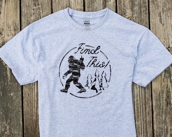 以下が含まれることがあります： ビッグフットの図柄がプリントされたグレーのTシャツ。ビッグフットは指を指しており、その周りに「Find This!」という文字が円形に書かれている。