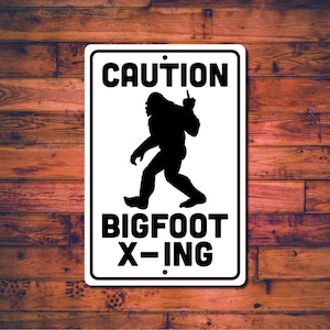 May include: A rectangular white metal sign with black text and a black silhouette of Bigfoot. The sign reads "CAUTION" at the top and "BIGFOOT X-ING" at the bottom. The Bigfoot figure is walking and giving the middle finger.