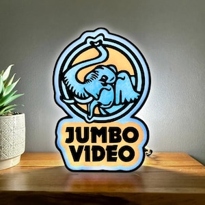 Peut inclure: Une enseigne rétroéclairée présentant un emblème circulaire avec une illustration d'éléphant bleu et le texte JUMBO VIDEO en lettres noires grasses. L'enseigne émet une lueur douce, soulignant les tons bleu clair et jaune sur les contours.
