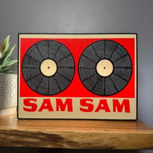 May include: A rectangular sign with two black vinyl records on a red background. The sign has a beige border and the text "SAM SAM" in red. The records have beige centres.