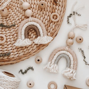 May include: Two macrame rainbow wall hangings in neutral colours, with cream fringe, are displayed on a white surface. Wooden beads and rings, along with a spool of twine and sprigs of greenery, are scattered around the rainbows. The rainbows have layers of beige, grey, and white.