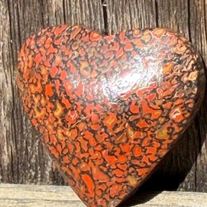 May include: A heart-shaped stone with a mosaic pattern in shades of orange, red, and black. The stone has a polished surface and is set against a weathered wooden background. The heart shape suggests a decorative or sentimental item.