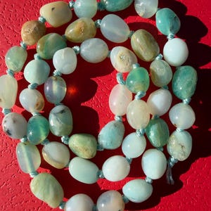 May include: A necklace featuring smooth, polished, tumbled stones in various shades of light green, teal, and off-white. The stones are strung together with a light blue cord, with individual knots tied between each bead to keep them separated.