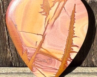 Puede incluir: Una piedra en forma de corazón con una superficie pulida. La piedra presenta una mezcla de tonos naranja, rosa y marrón con un patrón que se asemeja a un paisaje. El corazón mide aproximadamente 10 cm de ancho.