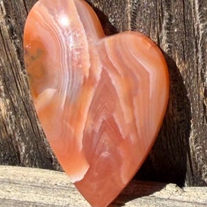 Puede incluir: Una piedra en forma de coraz&oacute;n con una superficie pulida. La piedra presenta bandas de color naranja, melocot&oacute;n y blanco, creando un efecto de capas. El coraz&oacute;n est&aacute; sobre una superficie de madera.