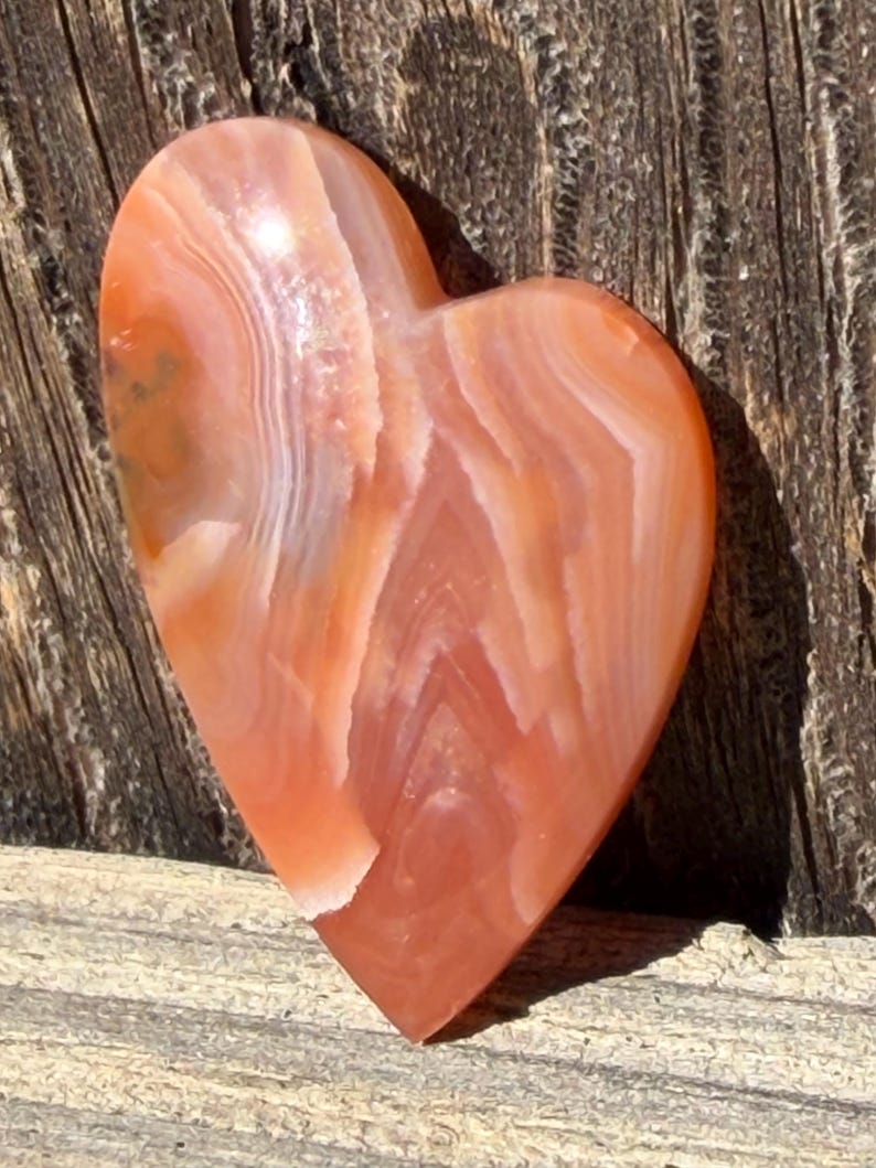Puede incluir: Una piedra en forma de coraz&oacute;n con una superficie pulida. La piedra presenta bandas de color naranja, melocot&oacute;n y blanco, creando un efecto de capas. El coraz&oacute;n est&aacute; sobre una superficie de madera.