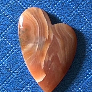 Puede incluir: Un colgante de piedra en forma de coraz&oacute;n con una superficie pulida. La piedra presenta patrones arremolinados en tonos marr&oacute;n, beige y crema. El colgante est&aacute; sobre un fondo azul texturizado.