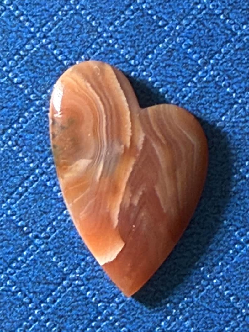 Puede incluir: Un colgante de piedra en forma de coraz&oacute;n con una superficie pulida. La piedra presenta patrones arremolinados en tonos marr&oacute;n, beige y crema. El colgante est&aacute; sobre un fondo azul texturizado.