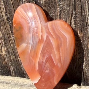 Puede incluir: Una piedra en forma de coraz&oacute;n con una superficie pulida. La piedra presenta bandas de tonos naranja, blanco y marr&oacute;n rojizo. El coraz&oacute;n se encuentra sobre un fondo de madera envejecida, resaltando los colores y texturas naturales.