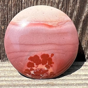May include: A round, polished stone with a layered appearance. The stone features bands of pink, red, and cream colors. A cluster of red-brown spots is visible near the bottom. The stone is set against a wooden background.