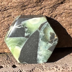 May include: A hexagonal gemstone with a mix of green and black colors. The stone has a polished surface and a unique pattern. The gemstone is set against a textured, brown background.