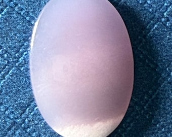 Puede incluir: Una piedra lisa, de forma ovalada, de color lavanda pálido, con aspecto esmerilado. El borde inferior tiene una sección blanca y opaca. La piedra está sobre un fondo azul oscuro texturizado.