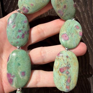 May include: A necklace featuring large, smooth, oval-shaped green stones with embedded magenta-colored inclusions. The stones are polished to a glossy finish and are connected by small, round, silver-toned metal beads along a thin string.