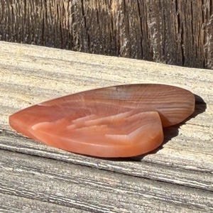 Puede incluir: Dos piedras pulidas en forma de l&aacute;grima, en tonos melocot&oacute;n y marr&oacute;n, sobre una superficie de madera envejecida. Las piedras tienen un acabado liso y brillante y muestran patrones de bandas naturales. La est&eacute;tica general es terrosa y org&aacute;nica.