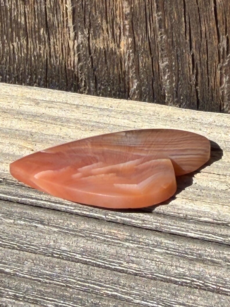 Puede incluir: Dos piedras pulidas en forma de l&aacute;grima, en tonos melocot&oacute;n y marr&oacute;n, sobre una superficie de madera envejecida. Las piedras tienen un acabado liso y brillante y muestran patrones de bandas naturales. La est&eacute;tica general es terrosa y org&aacute;nica.
