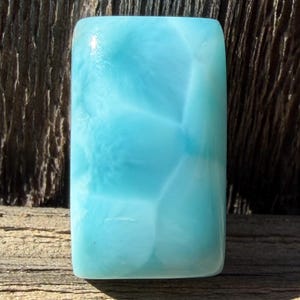 May include: A rectangular, polished gemstone with a light blue hue. The stone has a translucent quality, with internal patterns creating a marbled effect. The gemstone is set against a rustic wooden surface.
