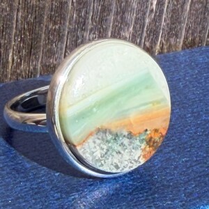 May include: A silver ring with a large, round stone. The stone features a landscape design with white, green, orange, and brown colors. The ring is set against a blue background.