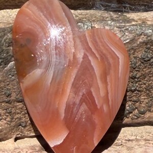 Puede incluir: Una piedra en forma de coraz&oacute;n con una superficie pulida. La piedra presenta bandas de tonos naranja, blanco y marr&oacute;n rojizo. El coraz&oacute;n es liso y parece estar hecho de &aacute;gata o un material similar. Es un objeto decorativo.