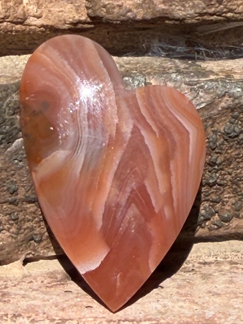 Puede incluir: Una piedra en forma de coraz&oacute;n con una superficie pulida. La piedra presenta bandas de tonos naranja, blanco y marr&oacute;n rojizo. El coraz&oacute;n es liso y parece estar hecho de &aacute;gata o un material similar. Es un objeto decorativo.