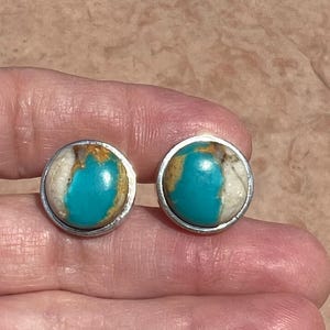 May include: A pair of silver earrings with turquoise stones set in a silver bezel. The turquoise stones have a blue and green color with white and brown veins.