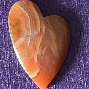 Puede incluir: Una piedra en forma de coraz&oacute;n con una superficie pulida. La piedra presenta una mezcla de tonos naranja, marr&oacute;n y blanco, con un patr&oacute;n en capas y arremolinado. El coraz&oacute;n est&aacute; sobre un fondo morado.