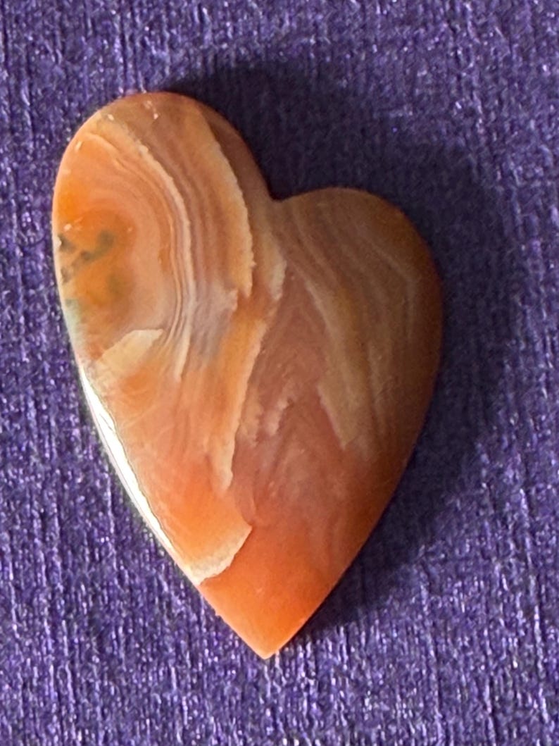 Puede incluir: Una piedra en forma de coraz&oacute;n con una superficie pulida. La piedra presenta una mezcla de tonos naranja, marr&oacute;n y blanco, con un patr&oacute;n en capas y arremolinado. El coraz&oacute;n est&aacute; sobre un fondo morado.