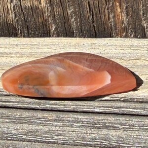 Puede incluir: Una piedra pulida de forma ovalada, con una mezcla de tonos naranja, marr&oacute;n y blanco. La superficie de la piedra tiene un aspecto natural veteado y se muestra sobre una superficie de madera envejecida.