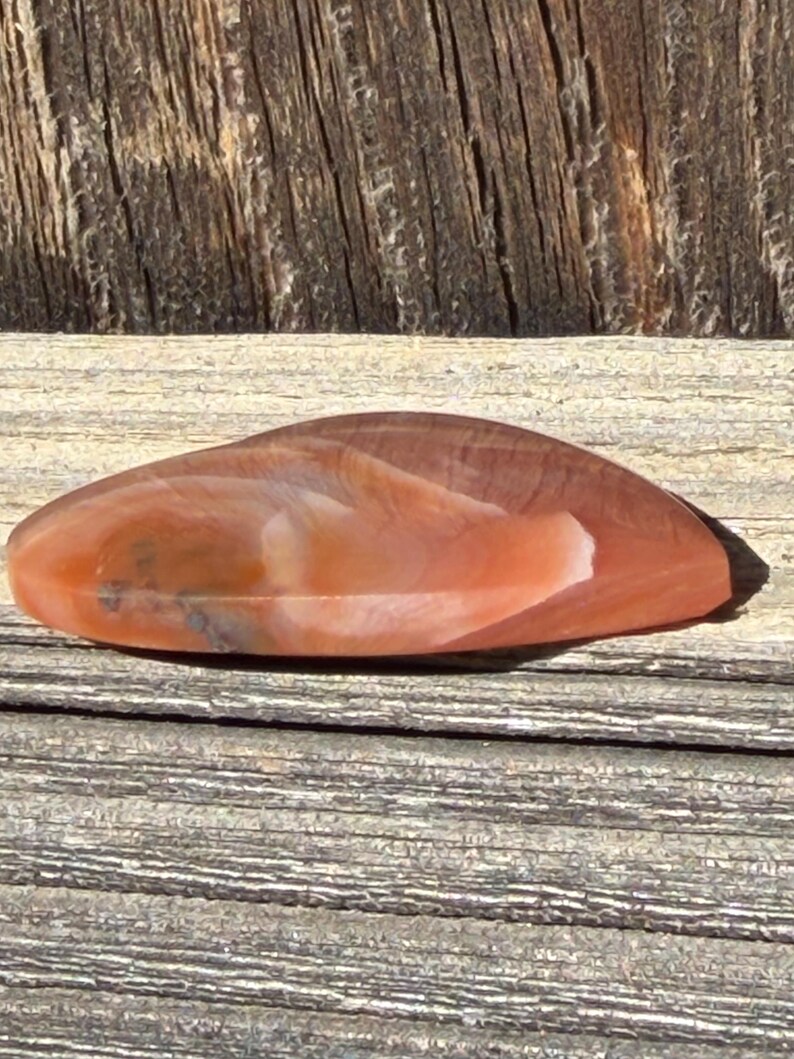 Puede incluir: Una piedra pulida de forma ovalada, con una mezcla de tonos naranja, marr&oacute;n y blanco. La superficie de la piedra tiene un aspecto natural veteado y se muestra sobre una superficie de madera envejecida.
