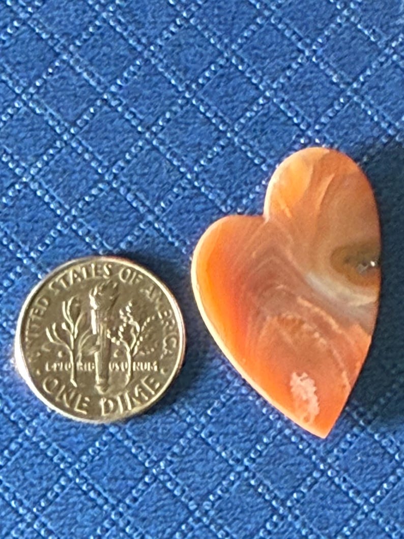 Puede incluir: Una gema en forma de coraz&oacute;n, de color naranja y blanco, junto a una moneda de diez centavos estadounidense. La gema tiene una superficie pulida con patrones naturales. La moneda muestra el perfil de Franklin D. Roosevelt y las palabras "ONE DIME". El fondo es una tela azul con estampado de diamantes.
