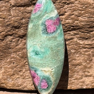 May include: A polished, elongated gemstone with a green base color and vibrant pink inclusions. The stone has a smooth surface and a natural, organic appearance. The pink inclusions are clustered, creating a unique pattern.