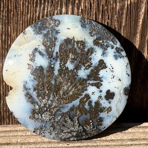 May include: A round, polished stone with a dendritic pattern. The stone features a light blue and white base with dark brown and silver-colored fern-like inclusions. The stone is set against a wooden background.