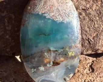 Puede incluir: Gema de forma ovalada con una llamativa mezcla de colores. La sección superior presenta un patrón texturizado en marrón claro y blanco, que transiciona a un vibrante centro turquesa y verde azulado. La parte inferior muestra una mezcla de tonos marrones, blancos y azules.