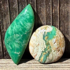 May include: Two polished gemstones. One is a vibrant green, elongated shape with swirling patterns. The other is a rounded stone with a mix of green, white, and tan colors. Both stones are displayed on a weathered wooden surface.