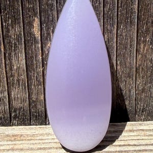 May include: A smooth, teardrop-shaped cabochon stone with a translucent lavender hue that transitions to a lighter, opaque white at the rounded base. The surface has a polished finish, reflecting light evenly across its curved, symmetrical form.
