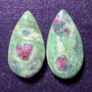 May include: Two teardrop-shaped gemstones. Each stone has a green base color with pink and blue inclusions. The pink inclusions are clustered in the center of each stone. The gemstones are set against a purple background.
