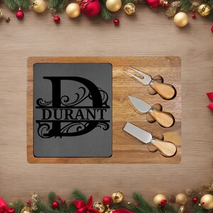 May include: A wooden cheese board with a slate insert engraved with the name "Durant" and a decorative initial. The board includes three cheese knives with wooden handles. Festive holiday decorations surround the board.