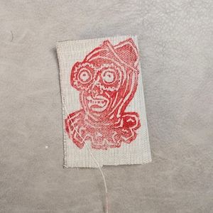 May include: A rectangular piece of light beige woven fabric featuring a red screen-printed graphic of a clown face with large circular eyes, a pointed hat, and a ruffled collar. A single loose white thread hangs from the bottom edge of the fabric.