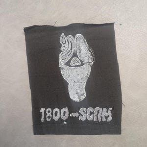 May include: A rectangular black fabric patch featuring a white screen-printed graphic of a stylized ghost mask with an elongated, melting appearance. Below the graphic, the text 1800-SCRM is printed in a bold, distressed white font.