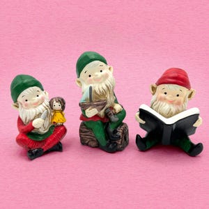 May include: Three small gnome figurines with white beards. The first gnome wears a green hat and red clothing while holding a small doll. The second gnome wears a green hat and green clothing, sitting on a log while holding a toy boat. The third gnome wears a red hat and green clothing while reading a black book.