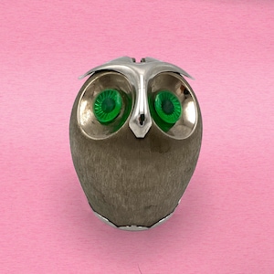 May include: A silver owl figurine with green glass eyes. The owl is made of metal and has a textured surface.