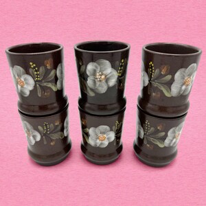 May include: A set of six dark brown ceramic tumblers arranged in three vertical stacks of two. Each tumbler features a hand-painted floral motif with white petals, yellow centers, and green leaves encircling the exterior of the cylindrical body.