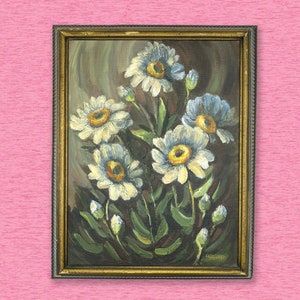 May include: A framed painting of a bouquet of white and blue flowers with yellow centers. The flowers are painted in a realistic style and are set against a dark brown background. The frame is gold and has a decorative border.
